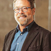 Studio-style portrait of a man wearing a blazer over a patterned button-up shirt against a neutral background.