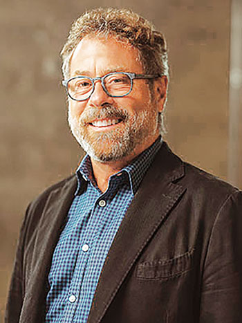 Studio-style portrait of a man wearing a blazer over a patterned button-up shirt against a neutral background.