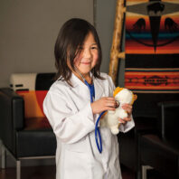 young girl wearing a white lab coat uses a toy stethoscope on a stuffed animal in an indoor seating area with patterned textiles in the background.