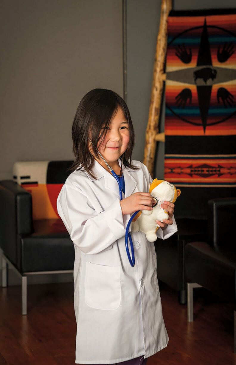 young girl wearing a white lab coat uses a toy stethoscope on a stuffed animal in an indoor seating area with patterned textiles in the background.