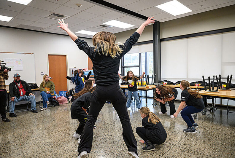 Group of children in a classroom space following a movement activity led by an adult with arms raised.