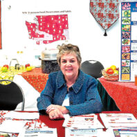 lt text: “Woman seated at an informational booth with pamphlets and food preservation materials, surrounded by posters and displays about home canning.