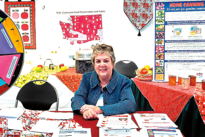 lt text: “Woman seated at an informational booth with pamphlets and food preservation materials, surrounded by posters and displays about home canning.