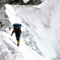 Mountaineer roped and walking along a narrow snow-covered ridge with steep ice walls on either side.