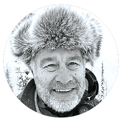 Circular black-and-white headshot of a man wearing a fur-lined hat, framed by a light border.