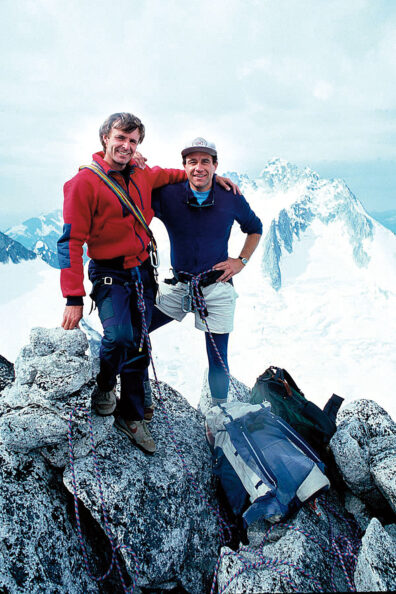 Two climbers standing on a rocky mountain summit with ropes and packs, snow-covered peaks stretching into the distance.