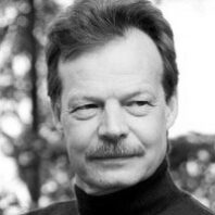 Black-and-white head-and-shoulders portrait of a man wearing a dark top, with trees softly blurred in the background.