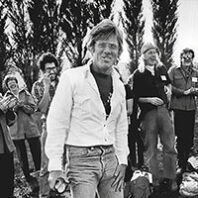 Black-and-white thumbnail image showing an adult standing outdoors in front of a small crowd, with trees in the background.