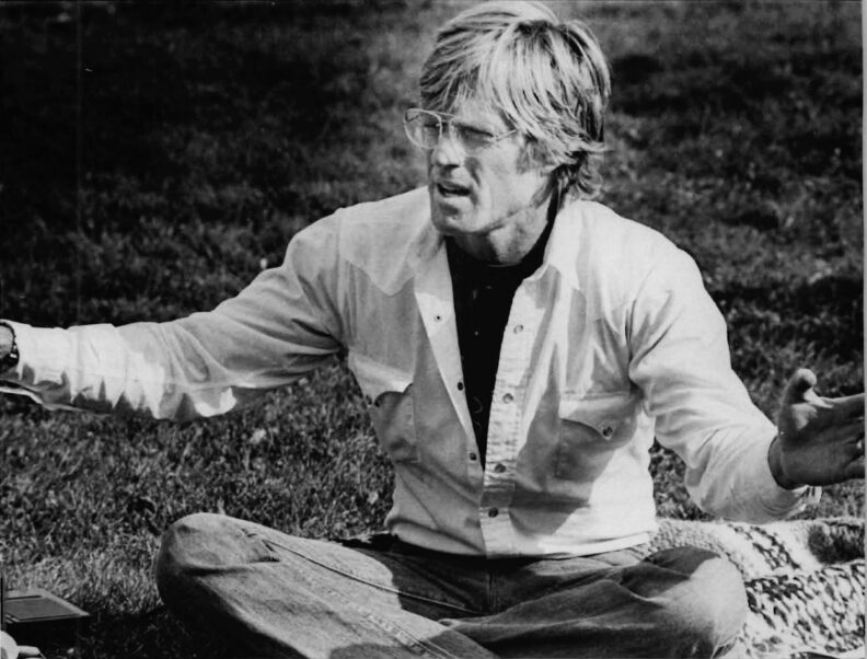 Black-and-white photograph of a man seated cross-legged on grass with arms extended, addressing a group gathered outdoors.