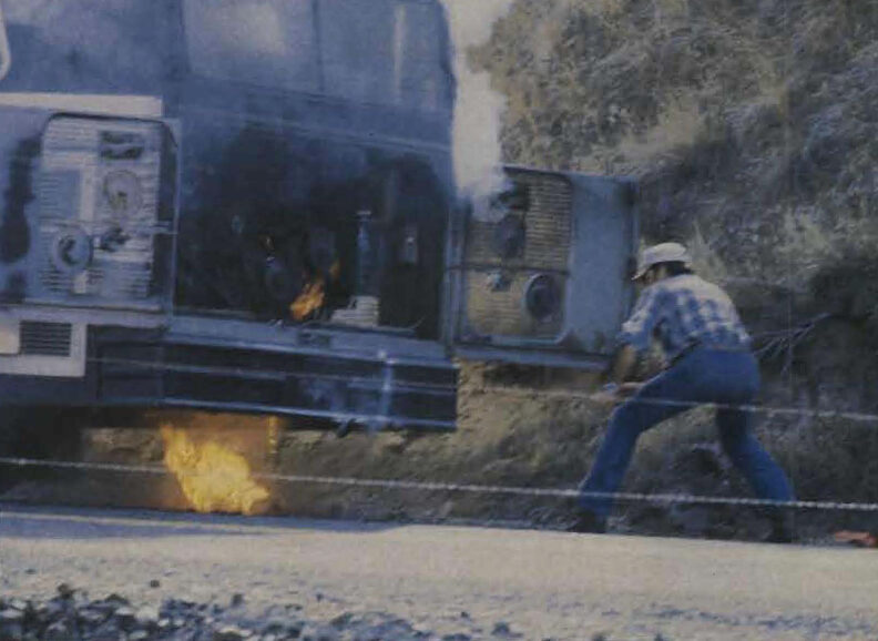 Rear of a bus stopped beside a rocky hillside, with flames visible underneath the engine area, while a person stands nearby pulling or handling an object connected to the bus.