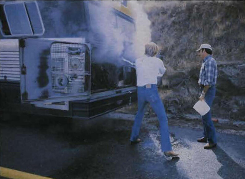 Two people standing behind a bus parked along a road, one facing the open engine compartment releasing smoke while another observes, with a hillside in the background.