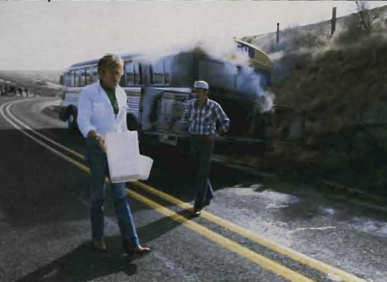 Two people standing behind a bus parked along a road, one facing the open engine compartment releasing smoke while another observes, with a hillside in the background.