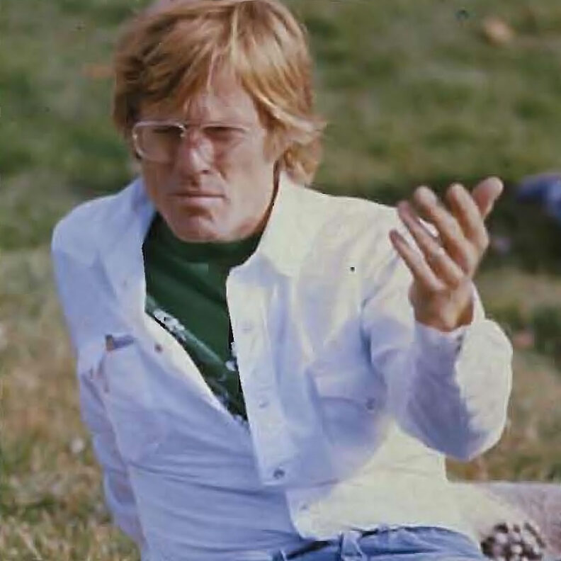 Adult seated on grass outdoors, wearing glasses and a light-colored shirt, with one hand raised and a blurred natural landscape in the background.