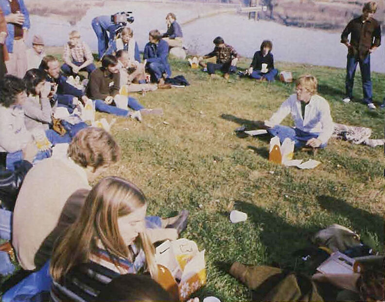 Group of people seated on grass in a loose circle outdoors, eating food and talking near a path and water, with bags and containers placed on the ground.