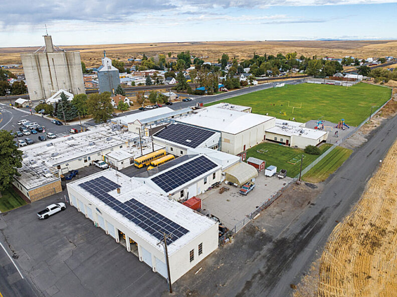 Aerial view of a school complex with solar panels installed on multiple rooftops, surrounded by streets, fields, and nearby buildings.