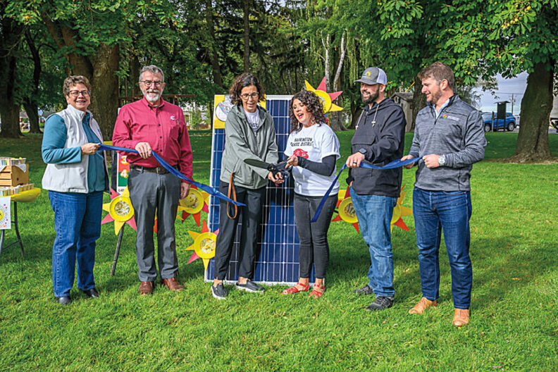 Group of people standing outdoors on grass holding a ribbon in front of a solar panel display during an event.