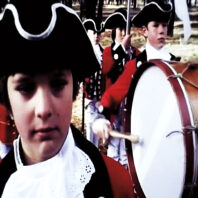 Historic-style reenactment scene showing people in colonial-era uniforms playing drums outdoors among trees.