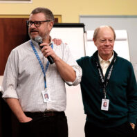 Two men standing indoors during a presentation, one holding a microphone, with presentation equipment in the background.