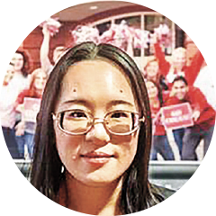 Circular headshot of a woman with long hair, with a blurred crowd scene and red signage in the background.