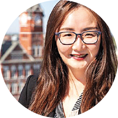 Circular headshot of a woman with long hair wearing a striped top, photographed outdoors with buildings behind.