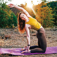Woman performing a side-bending yoga pose on a mat outdoors, photographed at sunset among trees.