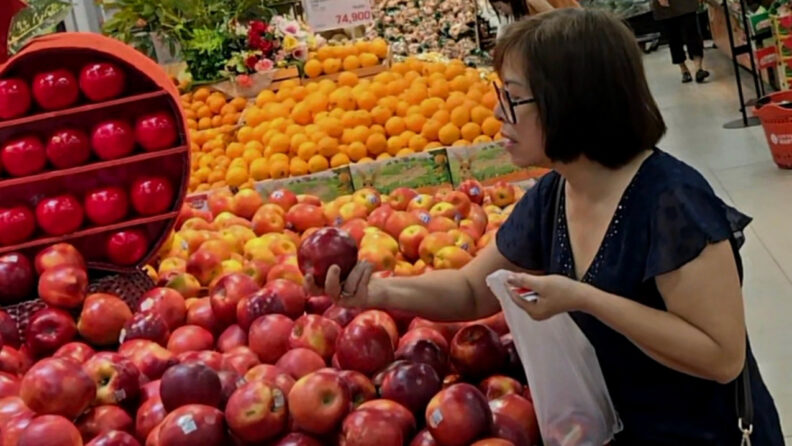 Person examining and selecting red apples at an indoor market display, holding a plastic bag while surrounded by piles of apples and oranges, with bright fruit stands and produce signage in the background.