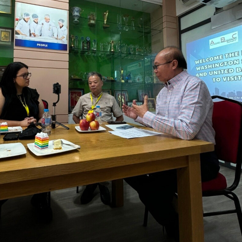 Three people seated around a table in an indoor meeting setting, engaged in conversation with a recording device on a tripod, plates of cake and apples on the table, and a presentation screen and trophy display visible in the background.