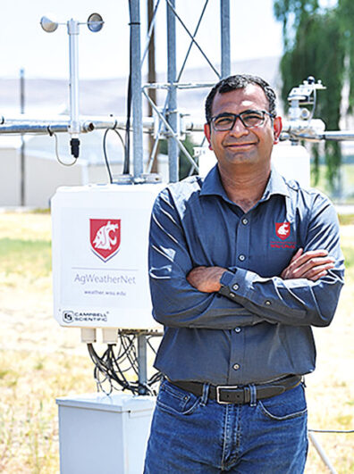 A man in glasses stands with arms crossed beside a weather monitoring station in an agricultural field. The station includes sensors and a white equipment box labeled AgWeatherNet.