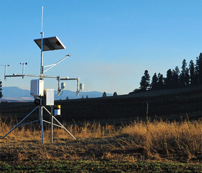 An automated weather station equipped with solar panels and meteorological sensors stands in a grassy field, with rolling hills and trees visible in the background.