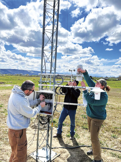 Three people install and maintain instruments on a tall weather monitoring tower in an open field under a sky filled with large clouds.