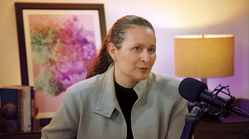 Woman speaking into a podcast microphone in a studio setting, with books, framed artwork, and a table lamp visible in the background.