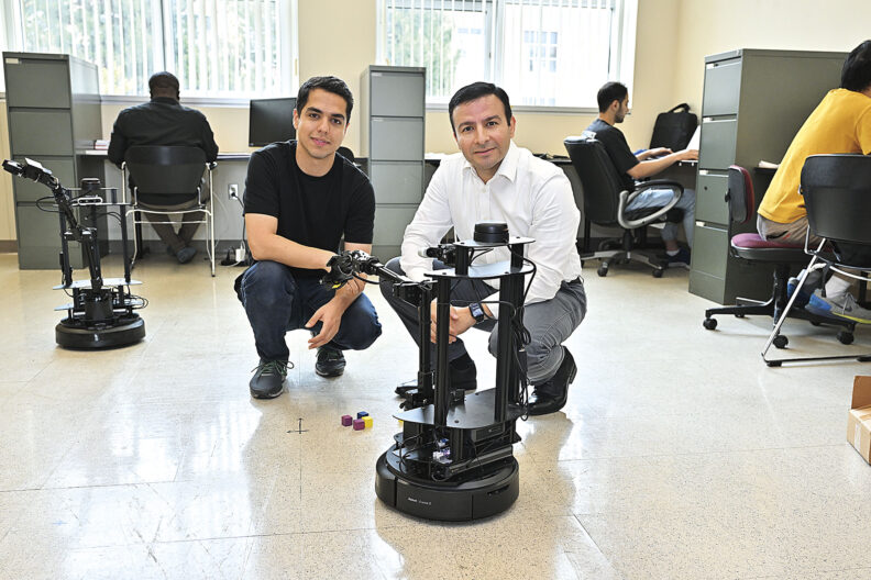 Two men crouch beside mobile robots in a robotics laboratory while other researchers work at computers in the background. One robot with a robotic arm is visible on the left, and a second autonomous platform is positioned in the foreground.