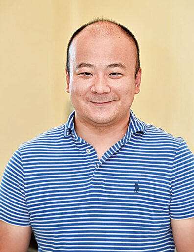 Portrait of a man wearing a blue-and-white striped polo shirt, standing indoors against a light-colored background.