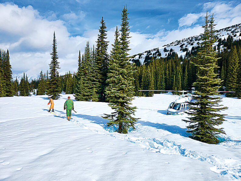 Two people walk across a snow-covered mountain landscape toward a helicopter parked among evergreen trees, with snow-covered slopes and a partly cloudy sky in the background.