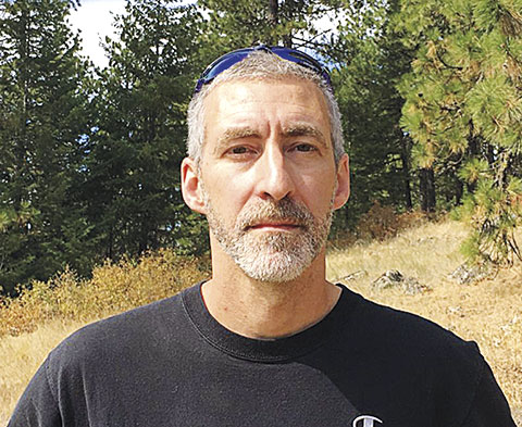 Portrait of a man with gray hair and gray beard standing outdoors in a forested area, wearing a black shirt with sunglasses resting on top of the head. Pine trees and dry grass are visible in the background.