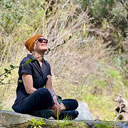Woman wearing sunglasses and a brimmed hat sits cross-legged on a rock in a natural setting with trees and brush in the background.