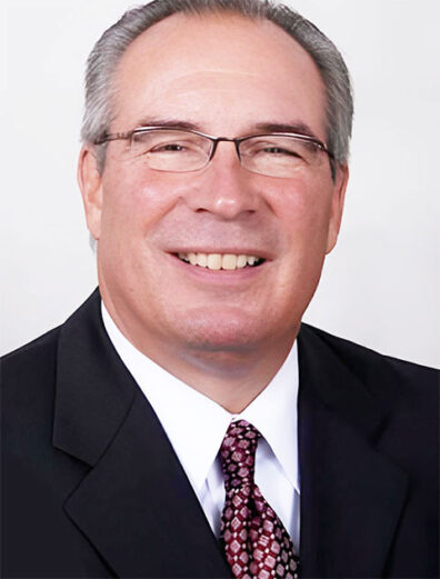Studio headshot of a man wearing glasses, a dark suit jacket, white shirt, and patterned tie against a light gray background.
