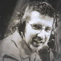 Black-and-white portrait of a young man wearing glasses and a broadcast headset with microphone, seated in front of a blurred crowd background.