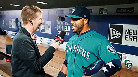 Sports reporter conducting an interview with a Seattle Mariners baseball player inside the team's dugout. The player wears a teal Mariners uniform.