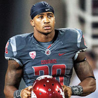 Close-up of a Washington State University football player in a gray uniform holding a crimson helmet while standing on the sideline.