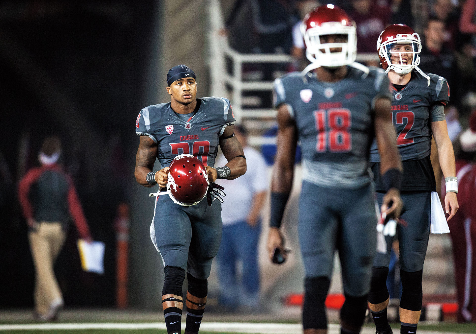 Washington State University football players in gray uniforms stand on the sideline during a game. A player carrying a crimson helmet is in focus while teammates and stadium activity appear in the background.