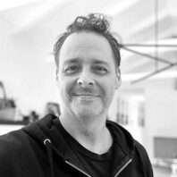 Black-and-white portrait of a man wearing a dark hoodie in an indoor setting, with a softly blurred background featuring office or studio furnishings.