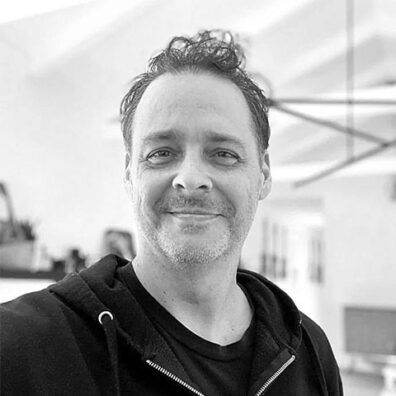 Black-and-white portrait of a man wearing a dark hoodie in an indoor setting, with a softly blurred background featuring office or studio furnishings.