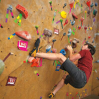 Man climbing an indoor bouldering wall covered with colorful handholds, footholds, and route markers. The climber is suspended on a steep section of the wall while navigating a climbing route.