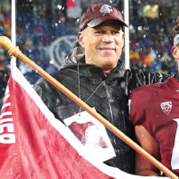 Washington State University football player wearing jersey number 47 stands with an arm around another man on a rain-soaked field while holding a WSU flag. Stadium lights and a crowd are visible in the background.
