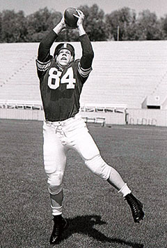Black-and-white photo of a football player wearing jersey number 84, leaping with both hands raised to catch a football on a stadium field.