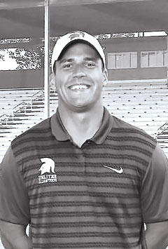 Black-and-white portrait of a young man wearing a team polo shirt and baseball cap, standing in front of stadium seating.
