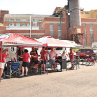 Washington State University fans gather under red-and-white Cougars-branded canopy tents at a tailgate area with trailers, RVs, and folding chairs set up near campus buildings.
