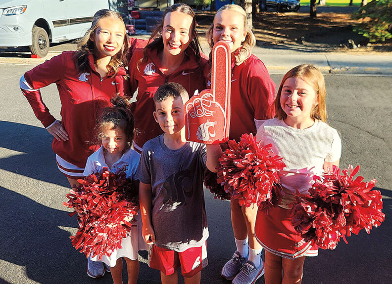 Three young women in Washington State University shirts and three children pose together outdoors, holding red pom-poms and a foam finger bearing the WSU logo.