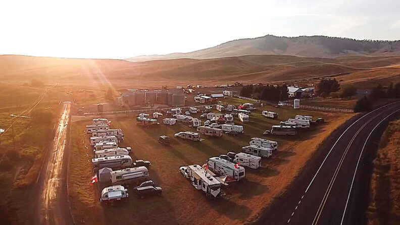 Aerial view of an RV tailgating area at sunset, with dozens of recreational vehicles parked in an open field beside a winding road and rolling hills.